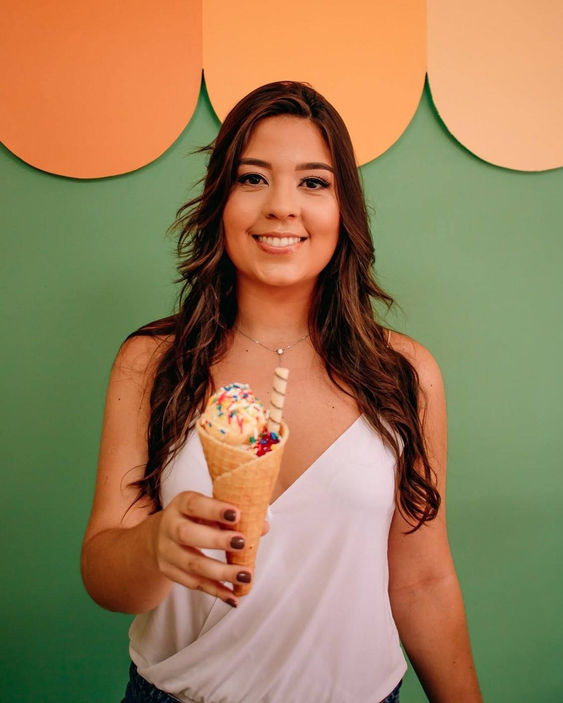 Elisa, neta da Tia Célia, em pé e sorrindo com um cascão de sorvete coberto de confeitos, diante da parede verde da loja.
