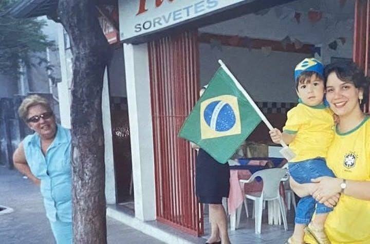 Tia Célia na calçada em frente à fachada antiga, ao lado da filha Luciana e da neta Elisa, ainda criança, vestida de verde e amarelo com uma bandeira do Brasil.