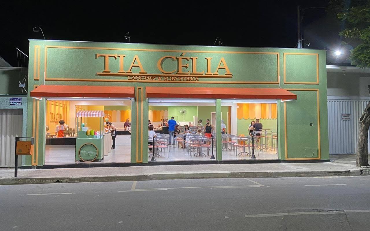 Fachada atual da Tia Célia Lanches & Sorveteria à noite, com letreiro dourado, toldos laranja e o salão iluminado cheio de clientes.