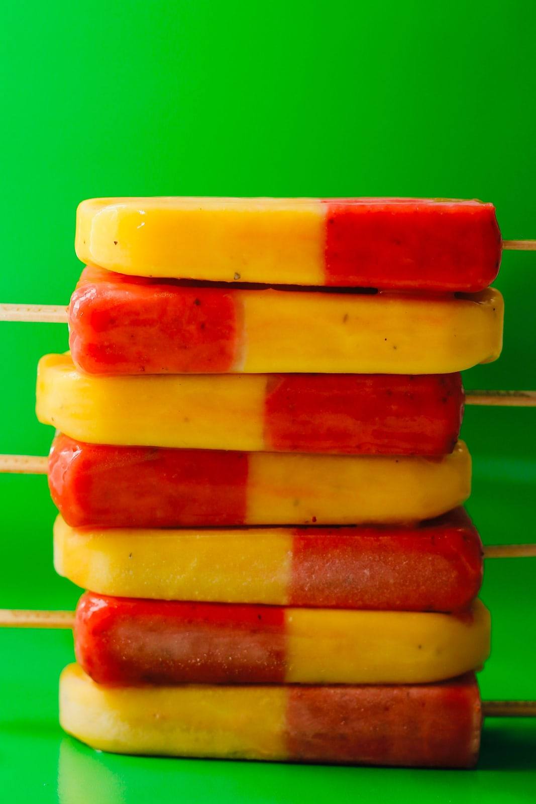 Pilha de picolés de morango natural com maracujá, metade vermelhos e metade amarelos, sobre fundo verde.