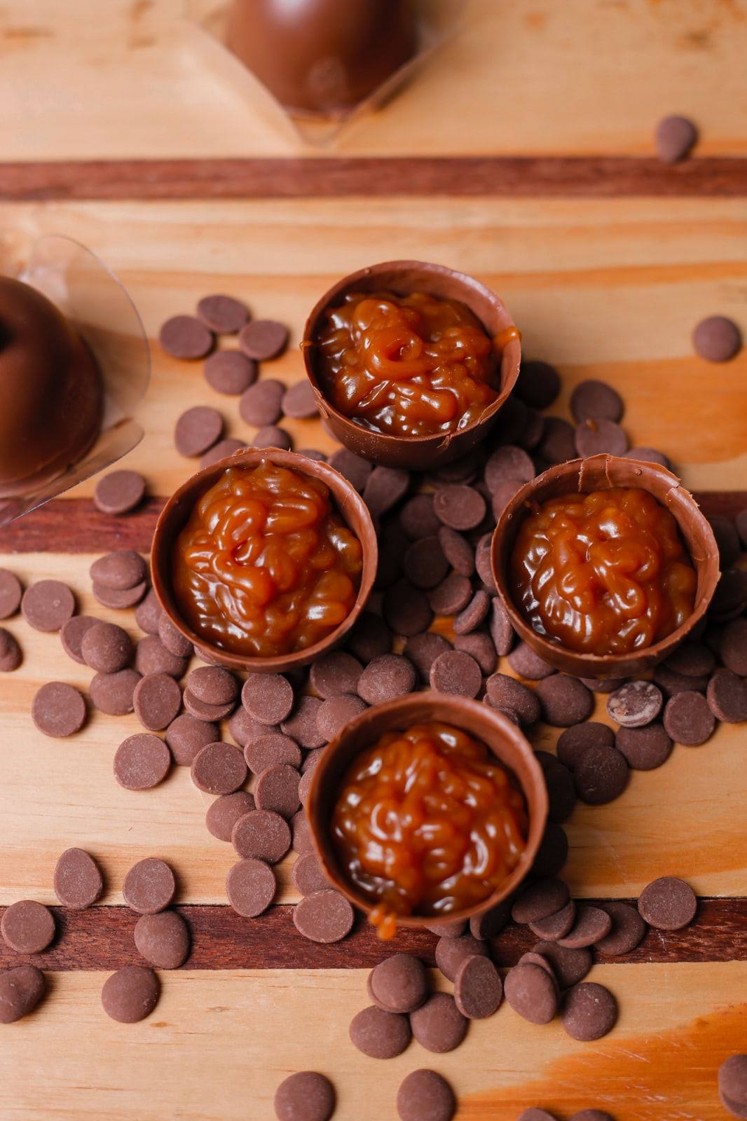 Calda de caramelo escorrendo para dentro de cascas de chocolate, cercadas de gotas de chocolate sobre tábua de madeira.