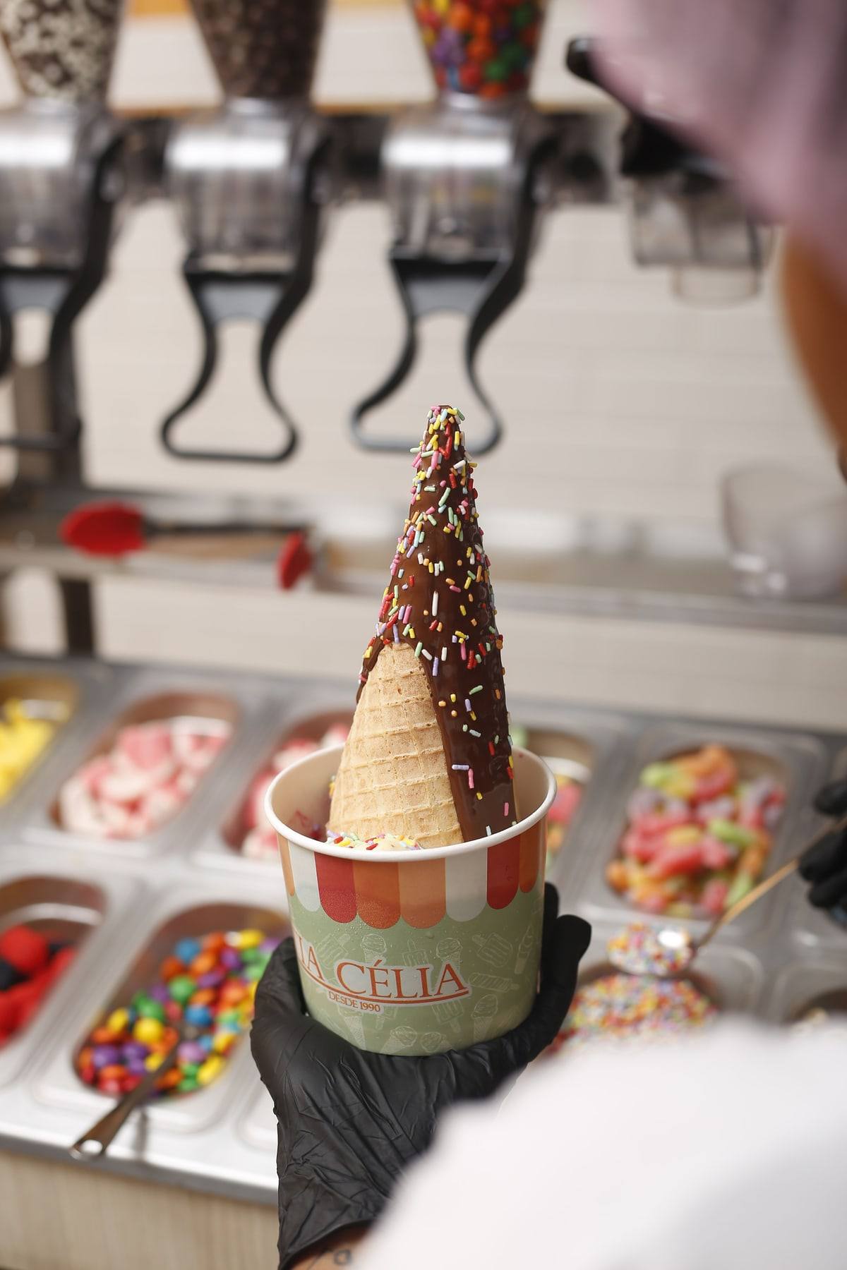 Cascão com borda de chocolate e granulado, servido num potinho da Tia Célia, diante das cubas de coberturas do self-service.